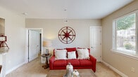 This single-floor residence offers a thoughtfully designed layout that balances comfort and functionality across several distinct living areas. The living room serves as the focal point of the home, featuring neutral beige walls, soft wall-to-wall carpeting, and a decorative compass rose wall accent that adds a nautical character to the space. The room accommodates a full-sized sofa and coffee table, with doorways leading to adjacent rooms and a closet for added storage.

A versatile den or office area connects to the outdoors through a glass door, flooding the space with natural light and providing views of a landscaped exterior. This room includes a built-in granite-topped desk flanked by warm wood cabinetry, making it well-suited for remote work or a secondary sitting area.

The bedroom is carpeted and comfortably sized, featuring a window with natural light and a wall-mounted mirror. The full bathroom is finished with wood-look flooring, a warm wood vanity with granite countertops, a large mirror, overhead lighting, and a shower-tub combination.

The kitchen is efficiently laid out with medium-toned wood cabinetry, granite countertops, wood-look flooring, and white appliances including a refrigerator, range, and built-in microwave. A dining area is visible adjacent to the kitchen, completing the home's functional flow.