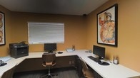 This single-floor business center is a thoughtfully designed workspace that caters to professionals seeking a functional and comfortable environment. The room is finished with warm, earthy amber-toned walls that lend a focused yet inviting atmosphere, complemented by dark carpeted flooring and a deep-toned ceiling that frames the space cohesively.

At the heart of the room is an L-shaped white work surface that wraps along two walls, providing generous desk space for multiple users simultaneously. Each workstation is equipped with a desktop computer monitor, keyboard, and mouse, making the space immediately ready for productivity. Ergonomic curved wooden chairs accompany each station, balancing style with comfort.

Natural light filters into the room through a window fitted with horizontal blinds, offering adjustable brightness to suit working conditions. A striking large-format framed art piece adorns one wall, adding a creative and polished touch to the otherwise utilitarian setup. A small organizer and desktop accessories are neatly arranged, reinforcing the workspace's practical character. This business center is a well-appointed and efficiently arranged space suited for tasks ranging from printing and browsing to focused remote work.