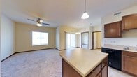 This single-floor residence features an open-concept layout that seamlessly connects the kitchen, dining, and living areas into one cohesive space. The kitchen includes a large central island with a light-toned countertop and dark wood cabinetry, providing ample workspace and storage. A pendant light fixture hangs above, offering focused illumination over the island.

The adjoining dining area is defined by an elegant chandelier, positioned near a sliding glass door that allows natural light to pour into the space while providing direct outdoor access. The living area extends further, featuring plush carpeting, a ceiling fan with integrated lighting, and a large window that brings in additional natural light.

The neutral wall tones and consistent trim work throughout the space create a cohesive, clean aesthetic. Dark wood accents in the cabinetry and door frames add contrast to the light carpet and countertops. Additional doorways visible from the main living area suggest access to private rooms and a bathroom. Overall, the layout is functional and well-lit, designed to maximize the open feel of the shared living spaces.