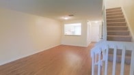 This two-story space features a clean, functional layout designed for everyday living. The main level showcases a spacious open living area finished with warm hardwood-style flooring that extends throughout, creating a cohesive and inviting atmosphere. Neutral cream-toned walls complement the light flooring, lending the space a bright and airy feel. A pass-through window connects the living area to the kitchen, offering both visual openness and practical convenience. The kitchen, visible through the pass-through, features white cabinetry and receives natural light from a window, enhancing its welcoming character. A hallway on the main level leads to additional rooms, including what appears to be a utility or storage area with a white door. A staircase with white railings, partially visible at the edge of the living area, provides access to the second floor, suggesting additional living or sleeping quarters above. Flush-mount ceiling lighting provides consistent illumination throughout. The overall design is straightforward and well-maintained, with clean lines and a neutral palette that offers a versatile foundation for a variety of uses.