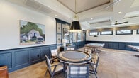Located at 1111 W Bay Ave in Barnegat, NJ, this single-floor game and recreation room offers a generously sized, thoughtfully designed space ideal for entertainment and leisure. The main game area is anchored by a dedicated poker table surrounded by plaid upholstered chairs, suspended beneath a striking black-and-brass pendant light fixture. Two billiards tables provide additional recreation space, while navy blue wainscoting and crisp white walls lend the room a sophisticated, nautical character. Recessed lighting and coffered ceiling details elevate the overall architectural design, while large multi-pane windows invite abundant natural light and outdoor views of the surrounding landscape. A cozy lounge nook, finished with richly textured wave-patterned wallcovering, features light blue upholstered armchairs, a warm wood dresser, and nautical-themed artwork, creating a comfortable retreat within the space. Hardwood flooring transitions to plush carpet throughout the main areas, adding warmth and comfort underfoot. Decorative framed artwork, including sailboat paintings, reinforces the coastal aesthetic. The overall ambiance strikes a balance between refined elegance and relaxed, inviting comfort, making this space well-suited for social gatherings and recreational use.