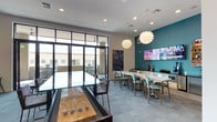 This single-floor amenity space is designed as a multifunctional social hub, combining entertainment, relaxation, and gathering areas within one open layout. The interior is anchored by a striking teal accent wall that contrasts with the neutral gray carpeting and white ceilings, creating a bold, contemporary aesthetic throughout.

A long white quartz bar-height counter runs along one side of the room, lined with patterned teal-and-wood bar stools. Above, two sculptural globe pendant lights serve as focal design elements. Mounted flat-screen televisions are positioned along the teal wall, making this area ideal for media viewing and socializing.

A dedicated game zone features a shuffleboard table and a sleek billiards table with high bar seating nearby. The lounge area offers a generously sized sectional sofa, accent chairs with gold frames, and eclectic art pieces — including decorative lattice panels and vibrant pop-art canvases — adding personality to the space.

Floor-to-ceiling black-framed glass doors flood the interior with natural light and open directly onto an outdoor patio furnished with high-top seating. This seamless indoor-outdoor connection, paired with the room's high ceilings and lively décor, gives the space an energetic yet welcoming ambiance.