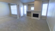 This single-story space centers around a generously sized living area finished with neutral-toned carpet that extends throughout the room. A standout feature is the white tile-surround fireplace, which anchors one wall and includes a built-in niche above the firebox, adding both function and visual interest. A stepped mantle and recessed window above the fireplace contribute to the room's architectural character.

The living area benefits from vaulted ceilings that enhance the sense of openness and volume. Multiple windows fitted with white horizontal blinds line the walls, allowing considerable natural light to fill the space while maintaining privacy. A glass-paneled door on one wall provides access to the exterior, further brightening the interior.

Decorative columns with detailed trim work frame a transitional area, subtly defining separate zones within the open layout without sacrificing the overall sense of spaciousness. The neutral beige walls and light carpet create a cohesive, understated palette throughout. Overall, the space presents a clean, open environment well-suited for comfortable living.