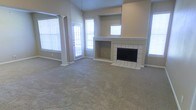 This single-story space centers around a generously sized living area finished with neutral-toned carpet that extends throughout the room. A standout feature is the white tile-surround fireplace, which anchors one wall and includes a built-in niche above the firebox, adding both function and visual interest. A stepped mantle and recessed window above the fireplace contribute to the room's architectural character.

The living area benefits from vaulted ceilings that enhance the sense of openness and volume. Multiple windows fitted with white horizontal blinds line the walls, allowing considerable natural light to fill the space while maintaining privacy. A glass-paneled door on one wall provides access to the exterior, further brightening the interior.

Decorative columns with detailed trim work frame a transitional area, subtly defining separate zones within the open layout without sacrificing the overall sense of spaciousness. The neutral beige walls and light carpet create a cohesive, understated palette throughout. Overall, the space presents a clean, open environment well-suited for comfortable living.