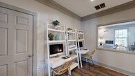 Located at 1001 Riverland Woods Pl in Charleston, SC, this thoughtfully designed business center offers a functional yet stylish environment for residents or guests. The single-room space is finished with warm wood-look flooring and crisp gray crown molding that frames the soft, neutral walls, lending a polished, cohesive aesthetic throughout.

The workstation area features two white ladder-style shelving units, each equipped with an iMac desktop computer and a woven rattan chair, providing comfortable and fully equipped spaces for productivity. A central shelving unit between the two desks includes a shared printer and additional display shelving adorned with potted greenery and decorative accents, keeping the space feeling fresh and inviting.

On the opposite side of the room, a mustard-yellow sofa offers a welcoming lounge area, complemented by a white side table and two framed art pieces displayed above. A sleek floor lamp adds a warm glow to this corner. A large interior window provides a view into an adjacent common area, enhancing the sense of openness, while a set of glass-paneled French doors offers a view into a fitness area beyond. The overall ambiance is bright, modern, and conducive to both focused work and relaxed downtime.