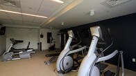 This single-floor fitness center offers a comprehensive workout environment designed to accommodate a variety of exercise routines. The space is anchored by a row of Matrix elliptical trainers positioned prominently at the forefront, providing ample space for cardiovascular training. Behind them, stationary bikes and a multi-station cable and strength training machine offer additional options for both cardio and resistance workouts. Free weights and a weight bench are also available, rounding out the strength training area. The room features a neutral-toned carpet flooring, a dropped ceiling with recessed linear and spot lighting, and a bold dark accent wall at the rear that lends the space a modern, energetic ambiance. The layout is thoughtfully arranged to allow comfortable movement between equipment. Overall, the fitness center presents a clean, well-maintained environment with quality commercial-grade equipment suited for a full-body workout experience.