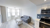 This single-floor space offers a clean and contemporary living environment defined by its minimalist aesthetic and thoughtful design. The main living area is bathed in natural light, courtesy of multiple windows fitted with horizontal blinds that allow for adjustable brightness throughout the day. Smooth, light-toned hardwood flooring runs throughout, complementing the crisp white walls and creating a warm yet modern atmosphere. The living space is well-proportioned, providing a comfortable setting for both relaxation and everyday living. A sleek gray sofa serves as the centerpiece of the room, paired with a minimalist gold-accented coffee table and a white accent chair positioned near the bay-style window corner — an ideal spot for reading or unwinding. A white dresser with gold hardware provides functional storage while contributing to the room's cohesive, contemporary palette. The overall ambiance is light, airy, and uncluttered, with a neutral color scheme that lends itself well to a variety of interior styling preferences. High, smooth ceilings and clean architectural lines reinforce the modern character of the space, making it feel open and inviting throughout.