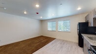 This single-floor unit offers a clean, contemporary layout designed for comfortable living. The kitchen is a standout feature, outfitted with dark wood-toned cabinetry, granite countertops, and a full suite of black appliances including a refrigerator, electric range, microwave, and dishwasher. Light wood-style luxury vinyl plank flooring runs throughout the kitchen and main living area, giving the space a bright, modern feel. Natural light flows in through multiple windows fitted with white horizontal blinds, providing both privacy and connection to the outdoors. The open living area transitions seamlessly from hard flooring to plush brown carpet, offering warmth and flexibility for furnishing arrangements. The carpeted bedroom is generously sized with two windows that invite natural light, white walls, and a paneled door leading to a closet. Recessed lighting throughout ensures consistent, even illumination across each room. The full bathroom is thoughtfully finished with a wood-toned vanity, granite countertop, large mirror, and a shower-tub combination, maintaining the cohesive aesthetic of the rest of the unit. Clean white walls and crisp white baseboards throughout create a neutral, move-in-ready canvas. The overall ambiance is bright, tidy, and functional.