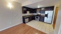 This single-floor space showcases a well-appointed kitchen that serves as a central feature of the layout. The kitchen is equipped with a full suite of stainless steel appliances, including a gas range with oven, built-in microwave, refrigerator with an in-door ice and water dispenser, and a dishwasher. Dark espresso-toned cabinetry lines the walls in an L-shaped configuration, providing generous storage both above and below the countertops. The countertops feature a dark granite or granite-style surface that complements the rich cabinet finish. A stainless steel sink is positioned along the lower cabinetry run, accompanied by a functional faucet. The kitchen floor is finished in light-toned ceramic tile, which transitions cleanly into warm hardwood-style flooring in the adjacent living area, creating a natural visual boundary between the two zones. The walls are painted in a neutral light gray tone, enhancing the brightness of the space. Overhead flush-mount lighting provides consistent illumination throughout the kitchen, while the open layout between the kitchen and living area contributes to an airy, connected feel throughout the space.