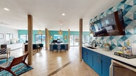 Located at 828 Orienta Ave in Altamonte Springs, Florida, this single-story community clubhouse showcases a bold, coastal-inspired design that is both visually striking and functionally diverse. The open floor plan connects multiple distinct zones, all unified by a cohesive palette of teal, blue, gold, and natural wood tones that give the space an energetic yet welcoming character.

The lounge area serves as a focal point, anchored by a dramatic large-scale teal octopus mural painted across a white brick accent wall. Mid-century modern seating, including teal sofas and wood-framed chairs, is arranged atop richly patterned blue area rugs, creating an inviting gathering space. Rope-wrapped columns add a nautical design element that carries throughout the clubhouse.

The main hall opens into a leasing or activity desk area, featuring sleek workstations framed by teal accent walls and rattan pendant lighting. Large windows and glass doors flood the space with natural light while offering direct views of the outdoor pool area. A wet bar station is outfitted with cobalt blue cabinetry, a white quartz countertop, under-counter refrigerator, and a bold geometric tile backsplash in teal and white with a wall-mounted television above. A billiards table anchors a dedicated game area, completing this multifaceted community hub.