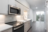 This single-floor unit showcases a thoughtfully designed open-concept layout with a consistent use of wood-look plank flooring throughout. The kitchen is well-appointed with white quartz countertops, flat-front cabinetry in a warm white finish with brushed gold hardware, a stainless steel undermount sink, and stainless steel appliances including a range, microwave, and dishwasher. A subway tile backsplash adds a clean, modern detail. The kitchen flows seamlessly into the living area, which is bright and open, benefiting from a large sliding glass door that leads to a private balcony overlooking lush, mature trees. The bedroom is generously proportioned, featuring a large window that brings in natural light and tree-lined views, alongside a sliding barn-style closet door for added style and functionality. The bathroom is finished in an all-white palette, featuring a tub-shower combination with tile surround, a vanity with white countertop, and warm-toned cabinetry consistent with the rest of the unit. Recessed lighting throughout, neutral white walls, and a clean, contemporary aesthetic create a cohesive and inviting atmosphere across all areas of the space.
