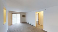 This single-floor unit features a 1-bedroom, 1-bathroom layout designed for comfortable everyday living. The main living area is carpeted and open, offering a generous sense of space enhanced by a large sliding glass door that floods the room with natural light and provides access to an outdoor balcony. A wall-mounted air conditioning unit ensures year-round climate comfort, while a baseboard heater along the exterior wall provides efficient supplemental warmth. The neutral wall tones and white ceilings contribute to a clean, bright atmosphere throughout. A doorway with warm wood-toned trim connects the living area to the bedroom, which continues the carpeted flooring and maintains a consistent, cohesive aesthetic. The straightforward layout promotes ease of movement between spaces, making the unit practical and easy to furnish. Overall, the space presents a simple, well-maintained interior that balances functionality with comfort, suitable for those seeking a manageable, low-maintenance living environment.