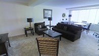 This single-floor space presents a thoughtfully arranged living area centered around a cohesive dark-toned furniture ensemble. The room features a large dark sofa adorned with decorative pillows, a matching loveseat, and a woven accent chair, all oriented around a dark wood coffee table that anchors the seating arrangement. End tables with matching dark-shaded lamps flank the seating area, providing ambient lighting and a sense of symmetry throughout the space.

The walls are finished in a soft, neutral tone that complements the carpeted flooring, contributing to a warm and inviting atmosphere. A framed piece of wall art adds a decorative focal point above the loveseat, while a wreath-style wall accent near the window adds a subtle decorative touch. Natural light filters into the space through a window dressed with horizontal blinds, brightening the room without overwhelming the neutral palette.

In the background, a dining area is visible, featuring dark wood chairs and a light-colored table, seamlessly extending the living space into a functional dining zone. The overall ambiance is cohesive, comfortable, and practically arranged for everyday living.