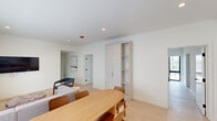 This single-floor residence offers a thoughtfully designed layout accommodating six bedrooms and five bathrooms within a generously sized floor plan. The interior embraces a modern aesthetic defined by crisp white walls, light-toned wide-plank flooring, and recessed lighting throughout. The open-concept living, dining, and kitchen area serves as the social heart of the home. The living room features large windows that invite natural light and provide outdoor views, complemented by a neutral sectional sofa and wall-mounted television. Adjacent to it, the dining area centers around a warm wood oval table with ample seating.

The kitchen is well-appointed with flat-front cabinetry in a light wood finish, stainless steel appliances including a gas range and full-size refrigerator, and an extended island with a stone countertop and bar seating — ideal for both cooking and casual dining.

The bedrooms maintain the home's clean, minimalist character, with the primary bedroom offering generous proportions and a floor-to-ceiling built-in wardrobe. The bathrooms feature large-format tile, glass-enclosed walk-in showers with matte black framing and built-in niches. A dedicated laundry room adds everyday convenience to this well-organized residence.