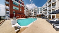 Located at 750 Thornton Way in Alexandria, Virginia, this outdoor amenity space offers a well-appointed pool and sundeck experience nestled within a landscaped courtyard. The centerpiece is a generously sized swimming pool with clear, blue water, framed by a clean concrete pool deck. An accessible pool lift is positioned poolside, underscoring the space's commitment to inclusive design. Adjacent to the main pool is a separate spa or wading area, providing additional relaxation options for residents.

The surrounding deck is outfitted with modern mesh chaise lounges and large patio umbrellas, creating comfortable shaded zones ideal for sunbathing or unwinding outdoors. A secondary elevated terrace area, enclosed by sleek glass railings, offers additional seating and views over the pool court. Decorative plantings and manicured landscaping add touches of greenery throughout.

The courtyard is enclosed on multiple sides by the building's multi-story facade, which blends classic red brick with white painted architectural detailing, balconies, and large windows — providing a sense of privacy while maintaining an open-air atmosphere. A pool house structure with a distinctive solar panel roof provides utility access. The entire setting benefits from abundant natural light, making it an inviting outdoor destination within the heart of Alexandria.