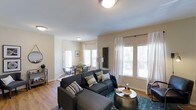 This single-floor residence offers an open, flowing layout that seamlessly connects the living and dining areas, creating a spacious and functional environment. The living area is anchored by large windows dressed with sheer white curtains and slatted blinds, allowing generous amounts of natural light to fill the space while maintaining privacy. The warm hardwood flooring runs throughout, adding continuity and a sense of warmth to the interior.

The living space comfortably accommodates a substantial sectional sofa paired with a stylish round metallic accent table, illustrating the room's generous proportions. Decorative elements such as patterned throw pillows and a teal area rug introduce color and texture against the neutral beige walls.

Adjacent to the living area, the dining space features a sleek glass-top table surrounded by metal chairs, positioned near its own set of windows that further enhance the bright, airy atmosphere. Flush-mount ceiling light fixtures provide consistent overhead illumination throughout both areas. The overall ambiance is light, modern, and inviting, with a clean, cohesive design aesthetic tying the entire space together.