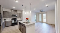 Located at 9140–9148 Ferguson Rd in Dallas, TX, this well-appointed two-bedroom, two-bathroom residence offers a thoughtfully designed single-floor layout with modern finishes throughout. The open-concept living and kitchen area serves as the heart of the home, anchored by a granite-topped island with a built-in sink and pendant lighting overhead. Taupe cabinetry, a tile backsplash, and black appliances complete the kitchen, while light wood-style flooring flows seamlessly into the living area, which features a ceiling fan and glass French doors that open to a private balcony. The primary bedroom is generously sized, finished with plush carpeting, a ceiling fan, and natural light from a window with blinds. It connects to a spacious walk-in closet outfitted with extensive wire shelving for ample storage. The en-suite primary bathroom features a dual-sink vanity with granite countertops, dark wood cabinetry, wood-style flooring, and a tiled shower. The second bedroom is bright and comfortable, also carpeted and bathed in natural light. The second bathroom mirrors the refined aesthetic with granite counters and a tiled tub-shower combination. Clean white walls and consistent finishes create a cohesive, modern ambiance throughout.