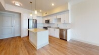 Located at 10 Westedge Street in Charleston, South Carolina, this single-floor residence offers a thoughtfully designed open-concept layout with modern finishes throughout. The kitchen serves as the centerpiece of the main living area, showcasing crisp white shaker cabinetry, a herringbone-patterned tile backsplash, and a full suite of stainless steel appliances including a range, microwave, refrigerator, and dishwasher. A center island with a light quartz countertop provides additional workspace and casual seating, while pendant lighting with gold-toned accents adds a refined touch. Light wood-style flooring flows seamlessly from the kitchen into the open living area, which benefits from generous natural light through large windows framing views of the surrounding Charleston cityscape. The bedroom features soft carpet flooring, a ceiling fan, and ample natural light from multiple windows. A spacious walk-in closet with wire shelving provides abundant storage. The bathroom is finished with clean white tile surrounding a soaking tub and shower combination, set atop light stone-look tile flooring. The overall aesthetic is clean, bright, and contemporary, reflecting the modern character of this well-positioned Charleston address.