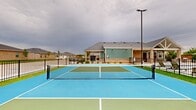 Located at 1800 NW 115th Terr in Oklahoma City, Oklahoma, this outdoor recreational facility centers on a well-appointed pickleball court finished in a striking two-tone blue and green surface with crisp white boundary lines and a regulation net. The court is framed by lush synthetic turf borders that provide a clean visual separation from the surrounding landscape. Tall sports lighting poles flank the court, ensuring visibility for extended play into the evening hours. The entire area is enclosed by a sturdy black wrought-iron perimeter fence, offering both security and an open, airy feel that allows views of the surrounding residential neighborhood. Adjacent to the court, a community swimming pool with lounge seating is visible, complemented by a single-story clubhouse building featuring a combination of brick and board-and-batten siding beneath a pitched shingle roof. A covered porch with ceiling fans extends from the clubhouse, creating a comfortable gathering space that overlooks the recreational amenities. Landscaped rock beds and plantings accent the edges of the facility, adding a polished finishing touch to the overall outdoor environment. Together, these amenities form a cohesive, well-designed recreational hub suited for active community living.