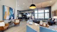 This single-floor communal space is thoughtfully designed to serve as a social and functional hub, blending modern aesthetics with practical amenities. The main lounge area greets visitors with an open layout anchored by a granite-topped reception counter finished in a cool blue-gray hue, complemented by light wood-look flooring that runs throughout the space. Recessed lighting and a striking black-and-gold pendant fixture illuminate the room, while large windows along one wall flood the interior with natural light and offer views of the surrounding exterior.

The lounge transitions seamlessly into a bar-height seating area equipped with mounted flat-screen televisions, ideal for casual gatherings. A dedicated conference room features a spacious rectangular table surrounded by navy upholstered chairs, a wall-mounted display screen, and an industrial-style circular chandelier, making it well-suited for meetings or group work sessions.

The fully equipped kitchen is a standout feature, offering expansive granite countertops, dark espresso cabinetry, stainless steel appliances including a French door refrigerator and dishwasher, and generous counter space running along both sides of the room. Natural light pours in through horizontal-blind-fitted windows. Throughout the space, curated artwork—including large abstract canvases and decorative carved wood panels—adds warmth and visual interest to the contemporary atmosphere.