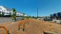 Located at 6026 Station Crossing Ave in Charlotte, NC, this outdoor dog park offers a well-appointed space for pets and their owners. The ground is fully covered in wood chip mulch, providing a safe and comfortable surface for dogs to run and play. A series of orange and green arch-style agility obstacles are arranged in a line through the center of the park, encouraging active engagement for dogs of various sizes. Additional enrichment features include scattered stepping stones and a bike-rack style leash post near the entrance. Sturdy black iron fencing encloses the entire perimeter, ensuring a secure environment, with a gated entry point for controlled access. Several metal benches are positioned throughout the space, offering resting spots for owners while they supervise their pets. Young pine trees and native shrubs are planted throughout, adding greenery and shade as they mature. A paved concrete pad is situated along one end, bordered by a large retaining wall that supports the adjacent multi-story residential building. The park enjoys open sky exposure, abundant natural light, and views of the surrounding residential community, creating a bright and welcoming outdoor amenity.