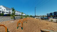 Located at 6026 Station Crossing Ave in Charlotte, NC, this outdoor dog park offers a well-appointed space for pets and their owners. The ground is fully covered in wood chip mulch, providing a safe and comfortable surface for dogs to run and play. A series of orange and green arch-style agility obstacles are arranged in a line through the center of the park, encouraging active engagement for dogs of various sizes. Additional enrichment features include scattered stepping stones and a bike-rack style leash post near the entrance. Sturdy black iron fencing encloses the entire perimeter, ensuring a secure environment, with a gated entry point for controlled access. Several metal benches are positioned throughout the space, offering resting spots for owners while they supervise their pets. Young pine trees and native shrubs are planted throughout, adding greenery and shade as they mature. A paved concrete pad is situated along one end, bordered by a large retaining wall that supports the adjacent multi-story residential building. The park enjoys open sky exposure, abundant natural light, and views of the surrounding residential community, creating a bright and welcoming outdoor amenity.