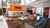 Located at 900 Radius Way in Newport News, Virginia, this polished two-story commercial space combines contemporary design with welcoming, functional interiors. The main level opens into a spacious lobby featuring large-format tile flooring, recessed lighting, and a prominent frosted glass installation that adds an artistic touch. Artwork adorns the walls, contributing to a gallery-like atmosphere throughout.

A standout feature of the first floor is the fireplace lounge, where a sleek, linear gas fireplace set against a bold orange accent wall serves as the focal point. Comfortable seating arrangements with striped and solid-upholstered chairs invite conversation and relaxation. Nearby, a well-appointed kitchen bar area features warm wood cabinetry, dark stone countertops, open shelving with barware, pendant lighting, and counter seating — ideal for casual gatherings.

Multiple lounge zones on the main level are furnished with modern seating and dining tables, all bathed in natural light from expansive windows offering views of the surrounding greenery and streetscape. Restroom facilities and additional amenity spaces are conveniently accessible off the main corridor.

The second floor features an open mezzanine with a striking steel-and-wood railing system overlooking the floor below. Large windows frame views of the neighboring urban landscape, flooding the upper level with abundant natural light and creating an airy, connected feel throughout the entire space.