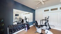 Located at 1019 Fox Hunt Ln in Raleigh, NC, this single-story space features a dedicated fitness room designed to support a full range of exercise routines. The room is finished with a bold navy blue accent wall that contrasts cleanly against white trim and walls, creating a modern and energizing atmosphere. A large wall-mounted mirror expands the visual depth of the space while allowing users to monitor their form during workouts. The flooring is finished in a neutral carpet, providing comfort underfoot during exercise. Overhead, a ceiling fan with integrated lighting ensures air circulation and a well-lit environment. Recessed lighting supplements the natural light that filters in through transom-style windows positioned near the ceiling. The fitness room is outfitted with a rack of dumbbells spanning a range of weights, a stability ball, treadmills, an elliptical trainer, and a recumbent bike, offering both cardio and strength training options. A wall-mounted television adds entertainment value during workouts. The layout is thoughtfully organized to maximize usable floor space while keeping equipment accessible and the overall environment open and functional.