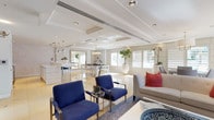 This refined single-floor space presents a seamlessly open layout with a polished contemporary aesthetic. The main lounge area is furnished with bold blue accent chairs with chrome frames, complemented by a neutral sofa with coral throw pillows, all anchored on a soft area rug over gleaming marble tile flooring. Tray ceilings with recessed lighting and integrated HVAC linear diffusers lend architectural depth, while plantation shutters along expansive windows allow abundant natural light to filter through.

A secondary lounge or meeting area features wall-mounted flat-screen TV, vibrant patterned accent chairs, and abstract artwork, with a glass-paned door providing connectivity to additional spaces. The dining area is highlighted by a modern rectangular table with upholstered chairs, a classic drum pendant chandelier overhead, and direct access to an outdoor area through French doors, offering an inviting connection to the exterior.

The kitchenette is sleekly outfitted with white cabinetry, a white quartz countertop, stainless steel appliances including a wine cooler, microwave, and refrigerator, alongside a wall-mounted TV and a textured botanical-patterned accent wall. A central white island with a mirrored chrome base anchors the kitchen zone, tying together the bright, cohesive palette that defines this well-appointed space.