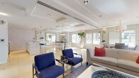 This refined single-floor space presents a seamlessly open layout with a polished contemporary aesthetic. The main lounge area is furnished with bold blue accent chairs with chrome frames, complemented by a neutral sofa with coral throw pillows, all anchored on a soft area rug over gleaming marble tile flooring. Tray ceilings with recessed lighting and integrated HVAC linear diffusers lend architectural depth, while plantation shutters along expansive windows allow abundant natural light to filter through.

A secondary lounge or meeting area features wall-mounted flat-screen TV, vibrant patterned accent chairs, and abstract artwork, with a glass-paned door providing connectivity to additional spaces. The dining area is highlighted by a modern rectangular table with upholstered chairs, a classic drum pendant chandelier overhead, and direct access to an outdoor area through French doors, offering an inviting connection to the exterior.

The kitchenette is sleekly outfitted with white cabinetry, a white quartz countertop, stainless steel appliances including a wine cooler, microwave, and refrigerator, alongside a wall-mounted TV and a textured botanical-patterned accent wall. A central white island with a mirrored chrome base anchors the kitchen zone, tying together the bright, cohesive palette that defines this well-appointed space.
