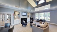 This elegantly appointed single-floor space exudes a refined, welcoming ambiance defined by its dramatic vaulted ceilings that draw the eye upward and create an impressive sense of openness and volume. Soft blue-grey walls complement the crisp white architectural details, including crown molding and built-in cabinetry that flank a classic fireplace serving as the room's focal point. A flat-screen television is mounted above the fireplace, seamlessly integrated into the built-in shelving units that display decorative accessories on either side. Natural light pours in generously through large windows and a striking arched transom window, illuminating the light-toned carpet and a carefully curated seating arrangement. Navy blue accent chairs, a neutral sofa, and a round coffee table create an inviting conversational grouping at the heart of the space. Toward the windows, a secondary seating area with upholstered chairs and a writing desk offers a more intimate setting with serene outdoor views of lush greenery. The overall design balances classic architectural charm with comfortable, functional furnishings, making this space well-suited for relaxation, social gatherings, or quiet activities.