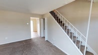 This two-story home offers a straightforward and functional layout spread across two levels. The main floor features an open living area with neutral-toned carpet, a front entry door with a transom window, and a staircase with white-painted railings leading to the upper level. Natural light filters in through blinds-fitted windows, creating a bright and welcoming atmosphere throughout.

The kitchen is equipped with warm honey-toned wood cabinetry, dark laminate countertops, and hardwood flooring. White appliances including a gas range, dishwasher, and refrigerator are neatly integrated, and ample upper and lower cabinet storage provides generous workspace for everyday use.

The upper floor hosts multiple carpeted bedrooms, each featuring neutral beige walls and windows with blinds that allow natural light to enter. The rooms offer versatile, open floor plans suitable for a variety of uses. A closet with bi-fold doors is accessible on the upper level, adding practical storage.

The full bathroom includes a vanity with a single-basin sink, warm wood cabinetry below, a large wall mirror, and a shower-tub combination with a curtain rod. The overall palette throughout the home is consistent, with soft beige tones and white trim lending a clean, cohesive aesthetic.