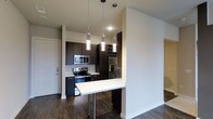 This single-floor residence showcases a contemporary design aesthetic defined by clean lines, dark contrasts, and modern finishes throughout. The open-concept kitchen serves as a central highlight, featuring rich dark espresso cabinetry that extends to the ceiling, providing ample storage while creating a bold visual statement. A suite of stainless steel appliances — including a French door refrigerator with ice dispenser, an electric range, and an over-the-range microwave — adds a polished, professional quality to the space. A crisp white peninsula countertop extends outward to form a breakfast bar with chrome-accented seating, offering a casual dining option while maintaining an open flow between the kitchen and the adjoining living areas. Three elegant cylindrical pendant lights hang in a row above the peninsula, casting warm, focused light that enhances the kitchen's modern ambiance. Dark hardwood-style flooring runs throughout, grounding the space and complementing the cabinetry's deep tones. The neutral, light-toned walls and ceilings provide a bright, airy contrast to the darker design elements, contributing to a well-balanced and inviting atmosphere. A dedicated entry door with modern hardware is visible, completing the functional and cohesive layout of this thoughtfully designed residence.
