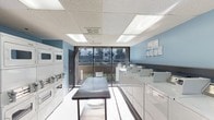 This single-floor laundry facility is designed with functionality and convenience in mind. The room features light blue walls that lend a clean, fresh aesthetic, complemented by white drop-ceiling panels with integrated fluorescent lighting that ensures bright, even illumination throughout. A row of stacked dryers lines one wall, while top-loading washers are arranged along the opposite side, providing ample capacity for multiple users. A sturdy central folding table offers generous workspace for sorting and folding laundry. Large floor-to-ceiling glass windows and a sliding door at the far end of the room flood the space with natural light and offer a view of an outdoor courtyard, enhancing the overall atmosphere. The light-toned flooring is easy to maintain and contributes to the room's clean appearance. Informational postings are displayed on the wall for user reference. The layout is open and thoughtfully organized, allowing for easy navigation between machines and the folding station, making this a practical and comfortable shared laundry space.
