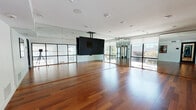 Located at 22121 Clarendon St in Los Angeles, this dedicated yoga and fitness studio offers a thoughtfully designed single-floor space ideal for movement, mindfulness, and group instruction. The main studio area is defined by rich, warm-toned hardwood flooring that extends throughout, creating an inviting and grounded atmosphere. Floor-to-ceiling mirrors line multiple walls, amplifying the sense of openness and allowing practitioners to monitor their form with ease. A ceiling-mounted flat-panel display serves as a central focal point, supporting instructor-led classes or guided sessions. Recessed ceiling lighting, combined with large black-framed glass walls and windows, floods the space with natural light and provides views of the surrounding greenery outside. The entry area transitions smoothly into the studio via sleek black-framed glass partitions and doors, maintaining an open and airy aesthetic throughout. A secondary corridor area features cork flooring, additional mirrored walls, and colorful artwork accents, adding a creative and welcoming touch. The clean white walls and modern architectural details throughout contribute to a calm, focused environment well-suited for yoga, dance, fitness training, or any wellness-oriented activity.