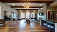 Located at 5150 Jack Byrnes Dr in Murfreesboro, Tennessee, this single-floor clubhouse facility masterfully combines rustic farmhouse aesthetics with contemporary design. The expansive main lounge serves as the centerpiece, showcasing rich reclaimed hardwood flooring laid in a distinctive pattern, soaring ceilings adorned with exposed wood beam details, and dramatic Edison-bulb chandeliers that cast a warm, inviting glow. Large French doors with transom windows line the walls, flooding the space with natural light and offering views to the outdoors. The lounge is furnished with a tufted leather sofa, velvet accent chairs, and a wall-mounted flat-screen TV above a fireplace, creating a sophisticated yet comfortable gathering area. A dedicated seating nook, framed by a sliding barn door, provides an intimate space for smaller meetings or consultations. Private offices with carpeted floors and built-in cabinetry are accessible via glass-paned French doors. The wellness wing includes a private tanning room and a dedicated relaxation suite equipped with an ergonomic zero-gravity lounge chair, acoustic wall panels, ambient LED lighting, and floor-to-ceiling drapery for a serene, spa-like experience. Throughout the space, thoughtful design details and high-end finishes create an atmosphere that is both functional and refined.