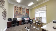 This single-floor community space serves as both a business center and casual lounge, thoughtfully designed to accommodate a variety of activities. The room is immediately distinguished by a large, colorful mural depicting a charming European streetscape, which anchors the space and adds a warm, artistic character to the environment.

Along one wall, a built-in workstation area features rich dark wood cabinetry and multiple computer terminals, providing residents with a dedicated space for productivity. The workstation area is finished with a light granite-style countertop, adding a refined touch to the functional setup.

At the center of the room, high-top bistro tables with sleek adjustable stools offer a casual gathering spot, decorated with small floral arrangements that bring a fresh, inviting feel. The patterned carpet in warm earth tones adds texture and visual interest underfoot.

Natural light fills the space through a large arched window with white trim, complemented by recessed fluorescent ceiling fixtures that ensure the room remains well-lit throughout the day. Crown molding along the ceiling lends an elegant, polished finish to the room.

A freestanding display rack near the window holds an extensive selection of magazines and books, making this corner an ideal spot for leisurely reading. The overall ambiance is warm, functional, and community-oriented.