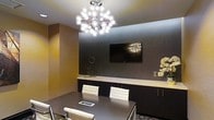 This stylish meeting room combines contemporary design with professional functionality. The space is anchored by a striking sputnik-style chandelier with multiple globe lights, serving as a bold focal point that draws the eye upward and adds a touch of modern elegance to the room. The ceiling is bright white, creating a clean contrast against the richly toned walls.

The walls feature a combination of warm golden-tan and deep charcoal-grey paneling, lending a refined, layered aesthetic to the interior. A large flat-screen television is mounted flush against the dark feature wall, making it ideal for presentations and collaborative work sessions.

A dark conference table takes center stage, surrounded by white upholstered chairs with chrome accents, balancing the darker tones in the room with crisp, clean contrast. Built-in dark cabinetry with a light countertop runs along one wall, providing ample storage and surface space. Decorative accents, including geometric sculptures and floral arrangements, add subtle personality without overwhelming the professional atmosphere. Overall, the space conveys a polished, business-ready ambiance well-suited for meetings and presentations.