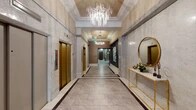 This sophisticated single-floor space presents a seamlessly connected series of upscale common areas designed to impress. The grand central corridor serves as the architectural centerpiece, showcasing floor-to-ceiling marble-clad walls, large-format tile flooring with contrasting border inlays, and a coffered ceiling adorned with a striking cascading crystal chandelier. Gold-toned elevator doors line one side of the hallway, while a gilded console table topped with a marble surface and an elegantly framed round mirror provide decorative accents. The corridor extends toward a glass-paneled entrance, allowing natural light to filter through and enhance the refined atmosphere.

Adjacent to the main corridor, a welcoming lounge area features rich leather seating, layered textiles, sheer floor-to-ceiling drapery, and a wall-mounted display screen beneath a circular brushed-gold pendant light. Artistic wall treatments and warm accent lighting contribute to the intimate ambiance.

A generously sized dining and gathering area accommodates multiple seating configurations, including a long communal wood table, individual dining tables with navy upholstered chairs, and bar-height seating along the perimeter. Circular pendant lighting, a tiled accent wall with pendant fixtures, and recessed tray ceilings complete the polished, contemporary aesthetic throughout.