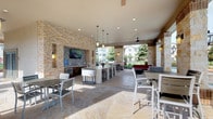 This single-floor space presents a thoughtfully curated blend of indoor sophistication and outdoor leisure. The interior dining area is anchored by a striking round marble-topped table in deep green tones, paired with woven rattan chairs atop a textured area rug over rich dark hardwood-style flooring. A large framed abstract artwork adorns the crisp white wall, adding an artistic focal point to the room. A sliding barn door with a weathered wood frame provides a charming transition to an adjoining space, where additional artwork and eclectic décor are visible through the opening.

The covered outdoor lounge area is equally impressive, featuring warm natural stone accent walls that lend a rustic yet refined character to the space. Generous seating is arranged throughout, including bar-height tables and chairs, as well as lounge furniture. Pendant and recessed lighting, along with ceiling fans, ensure comfort across all hours. A wall-mounted flat-screen television is set into the stone wall, making this an ideal space for entertainment. The open-air design invites abundant natural light and scenic views of surrounding greenery, creating an inviting and functional gathering environment suitable for both casual relaxation and social events.