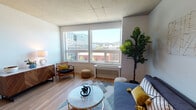 This contemporary urban residence offers a thoughtfully designed single-floor layout that blends modern industrial aesthetics with comfortable living. The open-concept kitchen is a standout feature, boasting dark cabinetry, stainless steel appliances including a gas range and built-in microwave, and a sleek white quartz waterfall island with an integrated sink. Pendant lighting and exposed concrete ceilings add an industrial character that carries throughout the space.

The living area is bright and welcoming, featuring large windows that frame sweeping city and hillside views, flooding the room with natural light and creating a strong connection to the urban surroundings. Warm wood-tone flooring runs throughout, complementing the light-toned walls.

The bedroom is a calm and restful retreat, accented by a grey feature wall and soft, neutral finishes. It connects seamlessly to the rest of the unit via the wood-floored hallway.

The bathroom is clean and functional, finished with grey subway tile in the shower-tub combination and crisp white fixtures. A stacked in-unit washer and dryer provides added everyday convenience. Overall, this space offers a cohesive, modern aesthetic with practical urban living in mind.