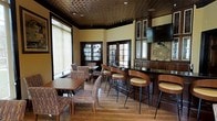 This single-floor space exudes a classic, upscale character defined by rich architectural details and thoughtful design. A striking embossed tin ceiling in a dark finish serves as a focal point overhead, adding texture and a sense of traditional elegance throughout the room. Warm wood tones are carried consistently across the bar cabinetry, bar stools, and trim work, creating a cohesive and inviting atmosphere.

The bar area features a sleek, dark countertop with stylish curved wooden bar stools that offer comfortable seating for guests. Upper cabinetry with glass-front panels provides display storage, while a wall-mounted television is integrated seamlessly into the bar setup. A decorative wall medallion adds an artistic touch near the bar.

Natural light filters into the space through multiple French-style windows and glass-paned doors fitted with roller shades, softening the overall ambiance. The lounge area is furnished with upholstered chairs and wooden tables, offering a more relaxed seating option. Hardwood flooring ties the entire space together, complementing the warm, rich palette. The result is a sophisticated environment suited for social gatherings and relaxed entertaining.