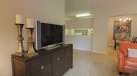 This single-floor residence features an open-concept design that connects the living area and kitchen in a cohesive, functional layout. The living area is finished with soft, neutral-toned carpet and painted walls in warm off-white hues, creating a welcoming and versatile space. A dark-finished media console with drawer storage serves as the focal point of the living area, providing both style and practicality for entertainment needs.

A pass-through opening connects the living space to the kitchen, enhancing the open feel while maintaining a defined separation between areas. The kitchen benefits from overhead fluorescent lighting that illuminates the workspace effectively, and appliances are visible through the pass-through, suggesting a well-equipped cooking area. The vaulted or raised ceiling detail adds a sense of vertical space and airiness to the interior.

The overall aesthetic throughout the space is understated and clean, relying on neutral finishes and simple architectural lines. The layout promotes ease of movement and a natural flow between living and culinary spaces, making it well-suited for everyday living.