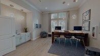 This single-floor amenity facility is thoughtfully designed to support both productivity and wellness. The coworking lounge is bright and inviting, featuring a large wooden communal desk equipped with desktop computers, geometric pendant lighting, and large windows that allow natural light to flood in while offering views of lush outdoor greenery. A built-in kitchenette with sage-green cabinetry and a sink adds functionality to the area, while a patterned accent armchair and framed coastal artwork contribute to a polished, contemporary aesthetic. Light wood-style flooring runs throughout, creating a cohesive visual flow.

Adjacent to the coworking space is a dedicated private office, appointed with a round conference table, upholstered chairs, bold navy patterned drapes, and a striking large-format bird photograph as a focal point. A separate formal conference room features a teal accent wall, a wall-mounted display screen, an oval meeting table surrounded by ergonomic chairs, and a sculptural pendant light fixture overhead, complemented by a curated gallery wall.

The expansive fitness center is a standout feature, divided into distinct cardio and strength training zones. The cardio area houses multiple treadmills and elliptical machines beneath wall-mounted televisions, with windows providing natural light and outdoor views. The strength zone offers a comprehensive array of Precor cable machines, weight benches, free weights, and resistance equipment, all supported by ceiling fans and mirrored walls that enhance the open feel of the space.