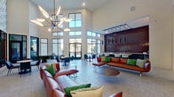 This single-floor amenity space makes a strong first impression with a grand entry corridor lined with bold black columns, textured decorative wall panels, and an intricate branch-patterned ceiling installation overhead. Light-toned tile flooring runs throughout, and black-framed glass double doors anchor the entry vestibule, accented by a sculptural chandelier.

The central lounge is the architectural centerpiece, featuring soaring double-height ceilings and an expansive wall of black-framed windows that flood the space with natural light and offer outdoor views. Rich cognac leather sofas with green accent pillows are arranged around low coffee tables, while a striking sculptural leaf chandelier commands attention from above. A bar counter nearby showcases a long white marble surface, matte black pendant lighting, and a dramatic floor-to-ceiling deep red tile backsplash. Framed artwork and a botanical-print accent wall with black-framed interior windows add visual depth to adjacent seating areas.

The games area includes a billiards table set against a ribbed gray accent wall with a mounted flat-screen TV, and a separate lounge hosts a shuffleboard table alongside sculptural green pod chairs and colorful modern artwork. Together, these zones create a cohesive, vibrant, and well-appointed communal environment.