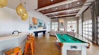 Located at 2855 Gulf to Bay Blvd in Clearwater, Florida, this single-story community clubhouse delivers a lively, coastal-inspired gathering space designed for recreation and relaxation. The open-plan interior is anchored by rich wood beam ceilings with recessed lighting, polished concrete floors, and soaring walls that maximize the sense of volume throughout.

The game and entertainment area features a full-size pool table and a shuffleboard table, complemented by a bar-height counter with vibrant orange metal stools — perfect for social gatherings. Large-scale, colorful beach-themed artwork lines the white shiplap accent walls, reinforcing the Florida coastal atmosphere.

The lounge zone offers a spacious sectional sofa arranged around a sculptural coffee table atop a bold geometric rug, alongside a fireplace and wall-mounted television. A wicker accent chair and warm pendant lighting add inviting texture to the space. Floor-to-ceiling glass-paned French doors and oversized windows flood the interior with natural light while framing views of the outdoor terrace and surrounding landscape.

A dedicated dining nook, bathed in light from tall transom windows and direct outdoor access, provides a quiet retreat within the space. A statement chandelier and curated coastal artwork throughout tie the design together, creating an energetic yet welcoming environment ideal for community use.