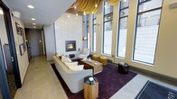 This contemporary lounge space makes an immediate impression with its dramatic double-height ceilings and an expansive wall of floor-to-ceiling windows that flood the interior with natural light and offer views to the exterior. The architectural design is bold and refined, highlighted by a striking sculptural ceiling installation featuring layered, wave-like golden forms that draw the eye upward and add artistic character to the space.

At the heart of the lounge sits a sleek, wall-mounted linear fireplace framed in crisp white stone, serving as a focal point that anchors the seating area. A generously sized neutral sofa paired with accent chairs, wooden side tables, and golden ottomans creates a warm and inviting arrangement set atop a rich, deep-purple area rug that contrasts beautifully with the large-format stone tile flooring.

Pendant lighting with globe bulbs is suspended at varying heights, adding warmth and visual depth to the already impressive volume of the room. The overall palette blends warm golds, soft neutrals, and deep purples to strike a balance between sophistication and comfort. This space is thoughtfully designed to serve as a welcoming, high-end communal or reception area with a distinctly modern aesthetic.