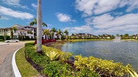 Located in Royal Palm Beach, Florida, this stunning lake entrance serves as a grand gateway to a well-appointed residential community. The approach is defined by a sweeping, herringbone-patterned brick driveway that curves gracefully alongside a serene community lake, creating an immediately impressive first impression.

The waterfront is a central highlight of this entrance, featuring a decorative fountain that rises from the calm lake surface, adding both visual interest and a sense of tranquility. The lake's edge is meticulously landscaped with vibrant tropical plantings, including tall royal palms, colorful bromeliads, manicured shrubs, and ground-level foliage in rich shades of green and red, all thriving under the abundant South Florida sunshine.

Flanking the driveway, the landscaping is carefully maintained, with neatly trimmed hedges, ornamental trees, and layered plantings that enhance the sense of arrival. The architecture visible throughout the community reflects a Mediterranean-influenced design, characterized by white stucco facades, dark wood accents, arched entryways, and multi-story buildings with covered balconies. Residences are set back thoughtfully, framed by mature palms that reinforce the lush, tropical character of the community. The overall ambiance is refined, welcoming, and evocative of classic South Florida resort-style living.