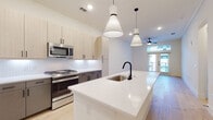 This contemporary single-floor residence showcases a clean, modern aesthetic defined by light hardwood flooring, crisp white walls, and thoughtfully selected finishes throughout. The heart of the home is an open-concept kitchen and living area, where a large central island with a sleek white quartz countertop and matte black faucet serves as the focal point. Striking pendant lights hang overhead, illuminating the space with both style and function. The kitchen is equipped with a full suite of stainless steel appliances, including a gas range, built-in microwave, and French door refrigerator. Two-toned cabinetry — light wood uppers paired with charcoal lower cabinets — adds visual depth, while a subway tile backsplash provides a subtle, timeless detail. The living area is bright and airy, featuring French doors that open to a balcony, inviting natural light deep into the room. Ceiling fans are installed throughout for added comfort. The bedroom is a serene, well-lit retreat with wide windows and continued hardwood flooring. The bathroom is appointed with large-format tile flooring, a dual-sink vanity with matte black fixtures, a generous mirror with Hollywood-style lighting, and a marble-surround soaking tub, delivering a spa-like experience.