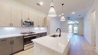 This contemporary single-floor residence showcases a clean, modern aesthetic defined by light hardwood flooring, crisp white walls, and thoughtfully selected finishes throughout. The heart of the home is an open-concept kitchen and living area, where a large central island with a sleek white quartz countertop and matte black faucet serves as the focal point. Striking pendant lights hang overhead, illuminating the space with both style and function. The kitchen is equipped with a full suite of stainless steel appliances, including a gas range, built-in microwave, and French door refrigerator. Two-toned cabinetry — light wood uppers paired with charcoal lower cabinets — adds visual depth, while a subway tile backsplash provides a subtle, timeless detail. The living area is bright and airy, featuring French doors that open to a balcony, inviting natural light deep into the room. Ceiling fans are installed throughout for added comfort. The bedroom is a serene, well-lit retreat with wide windows and continued hardwood flooring. The bathroom is appointed with large-format tile flooring, a dual-sink vanity with matte black fixtures, a generous mirror with Hollywood-style lighting, and a marble-surround soaking tub, delivering a spa-like experience.